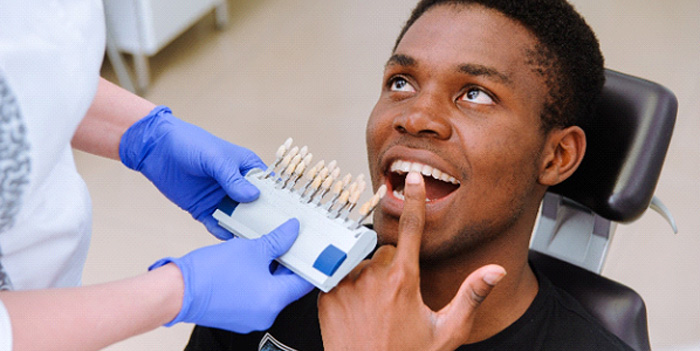 Man in dental chair pointing to teeth with shade guide held to his smile